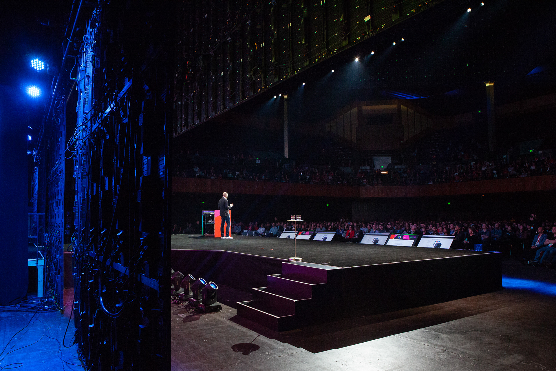 View from backstage wing of speaker addressing a full arena of thousands of attendees